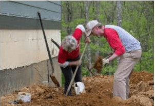 Two volunteers dig beside a house during an outdoor community project.