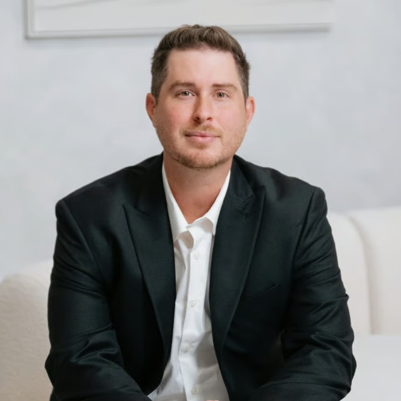 Man in a dark blazer and white shirt seated against a bright interior background.