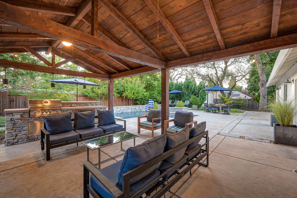 Covered patio with wood-beam ceiling, lounge seating, outdoor kitchen, and swimming pool.