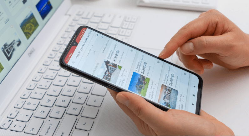 Hands browse home listings on a smartphone above a laptop keyboard.