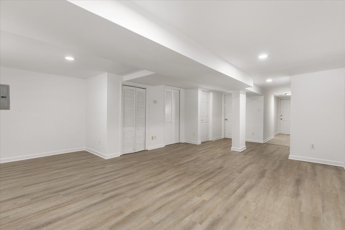 Unfurnished basement room with wood-look flooring, louvered closet doors, and a tiled hallway.