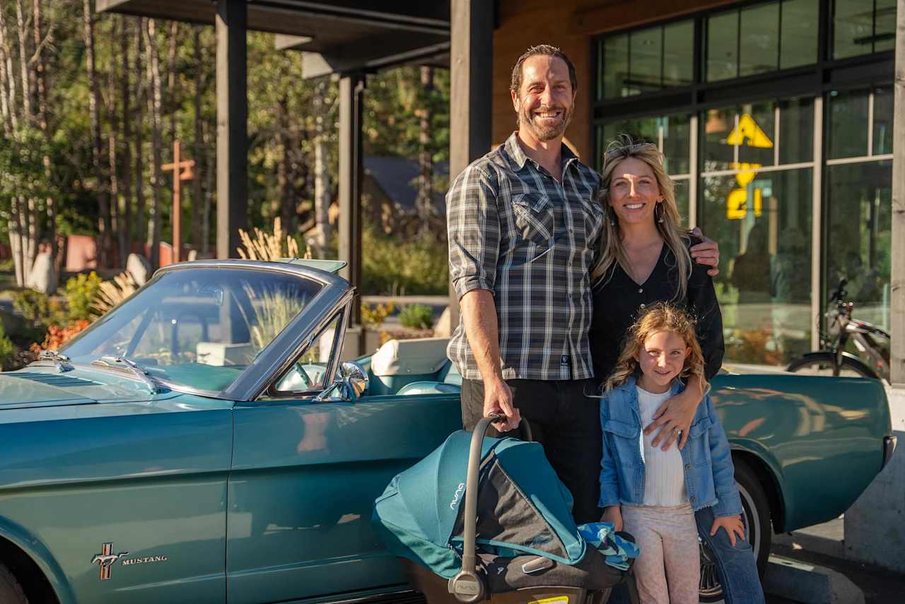 A family poses beside a teal vintage Ford Mustang outside a glass-fronted building.