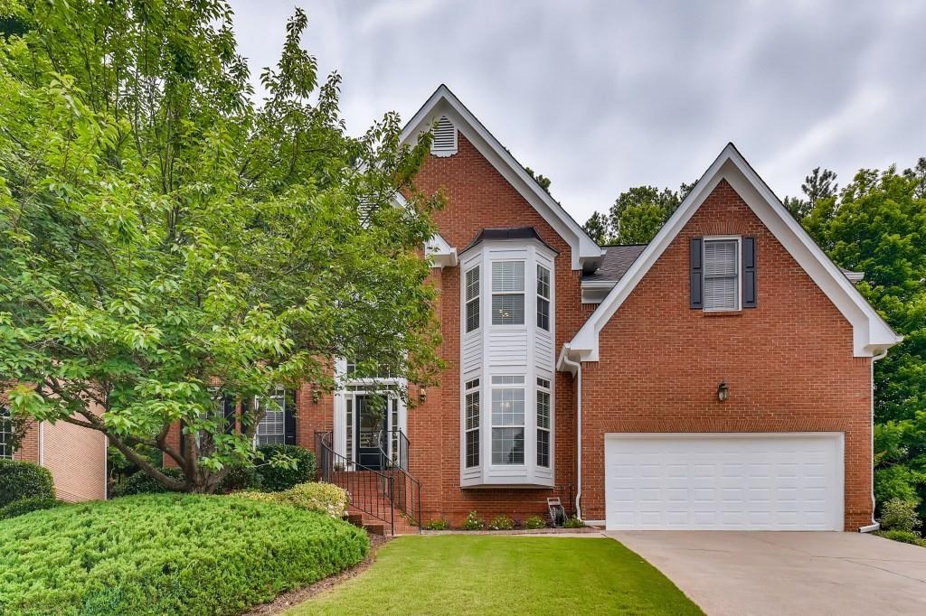 Red brick house with a white bay window, attached garage, lawn, and mature landscaping.