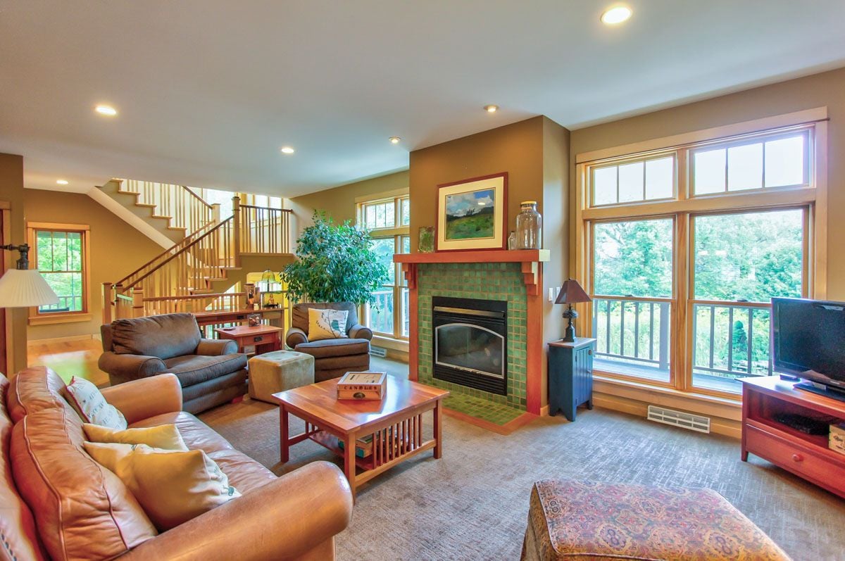 Living room with leather sofas, a green-tiled fireplace, and large windows overlooking trees.