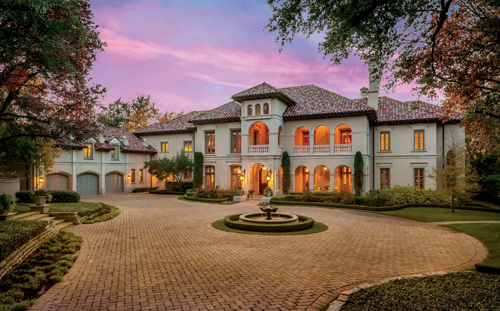 Large Mediterranean-style estate with illuminated arches, circular fountain, and brick motor court at dusk.