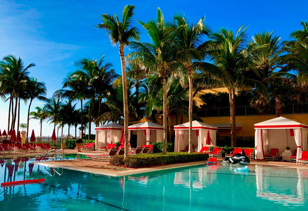 Resort pool with palm trees, red lounge chairs, white cabanas, and an inflatable orca.