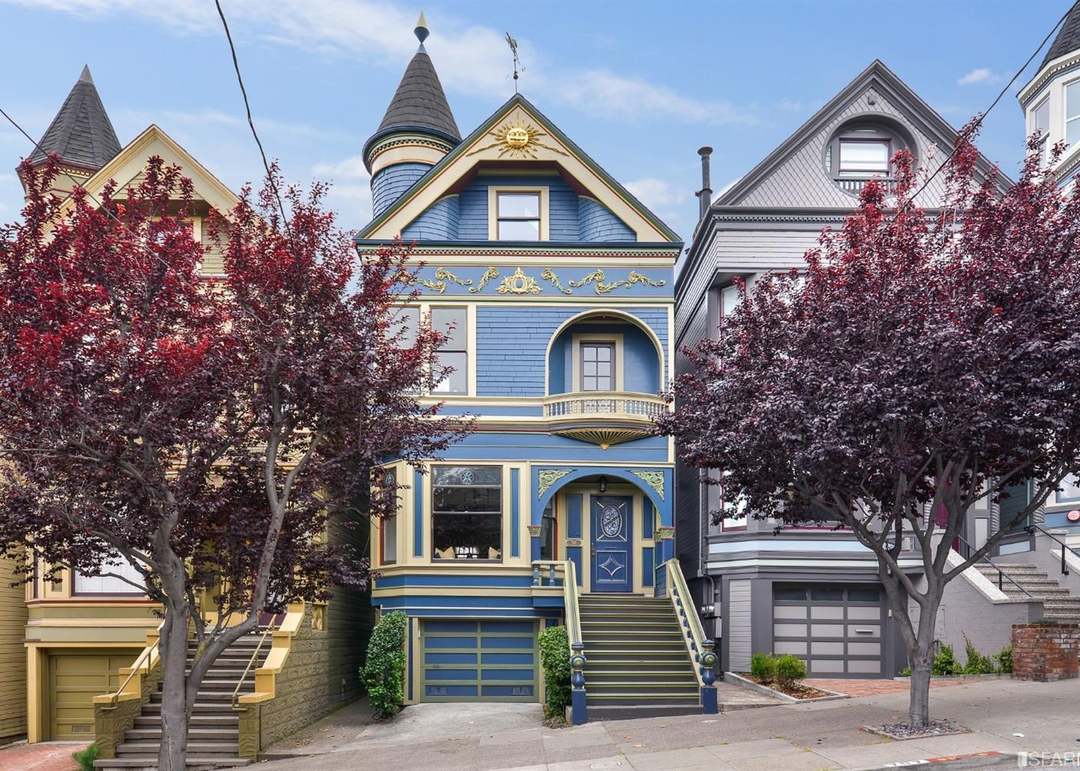 Blue Victorian home with an ornate turret and cream trim, framed by purple-leaved street trees.