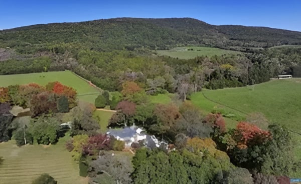Aerial countryside with green fields, autumn trees, scattered buildings, and a wooded ridge.