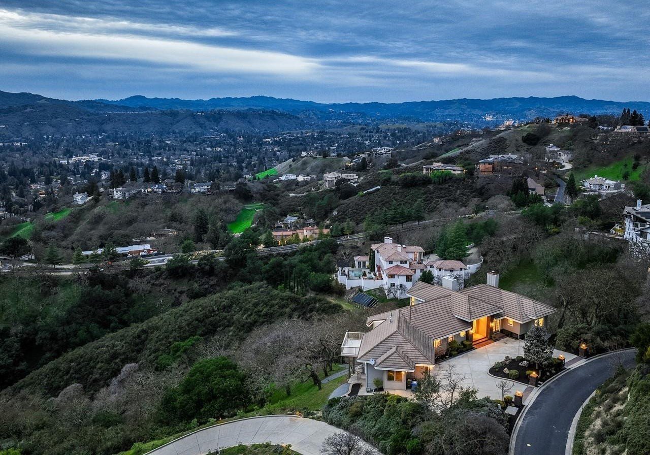 Hillside homes overlook a valley of houses and wooded hills, with warm lights glowing in the foreground.