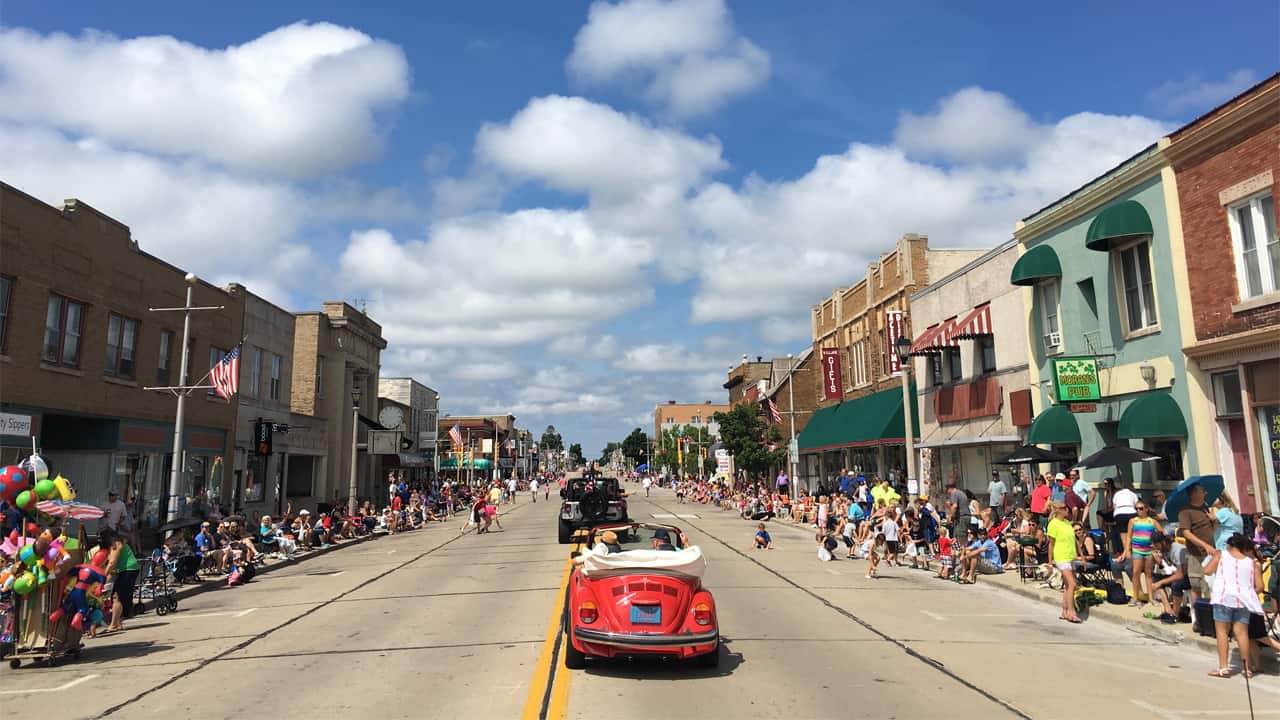 Crowds line both sides of downtown South Milwaukee as cars drive through a community parade on a sunny day.
