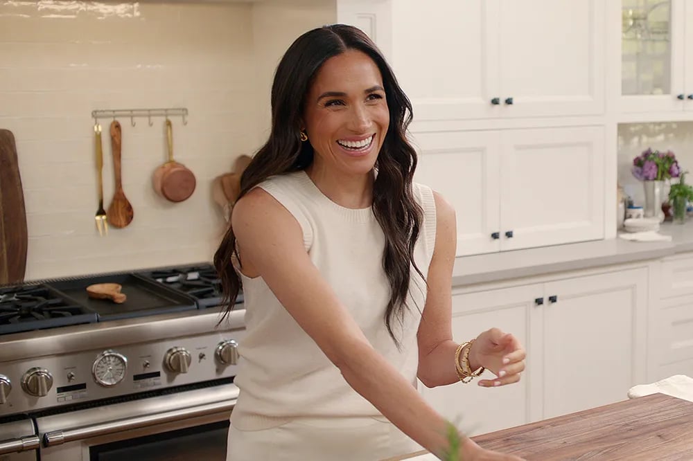 Meghan Markle smiles in a sunlit Montecito kitchen featuring white cabinetry, artisanal copper cookware, and rustic-modern finishes—reflecting the refined lifestyle aesthetic influencing high-end buyers. Brendan Brown Real Estate clients increasingly reference this visual language when seeking emotionally resonant luxury estates in Montecito, Beverly Hills, and Brentwood.