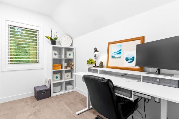 Furnished home office with a long white desk, black chair, large monitor, and window with white blinds.