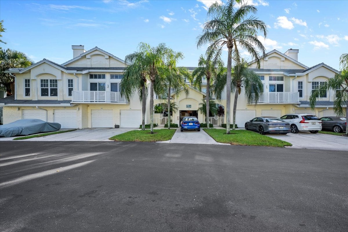 Cream-colored condominium building with garage doors, balconies, a central entrance, and palm trees.