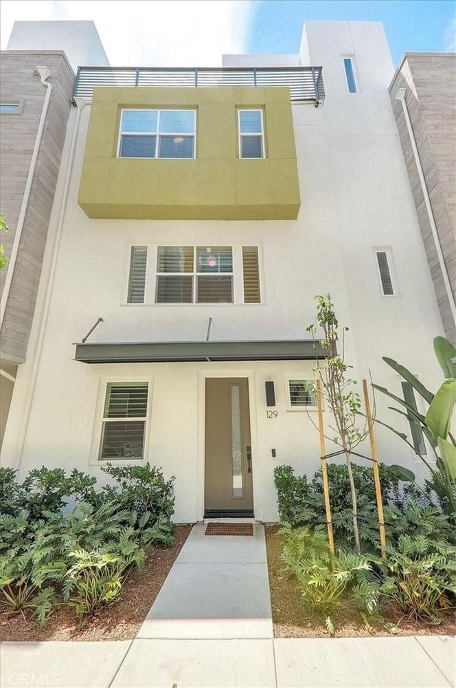  A narrow, three-story modern townhome with a light green accent box on the second floor and a small, landscaped front yard.