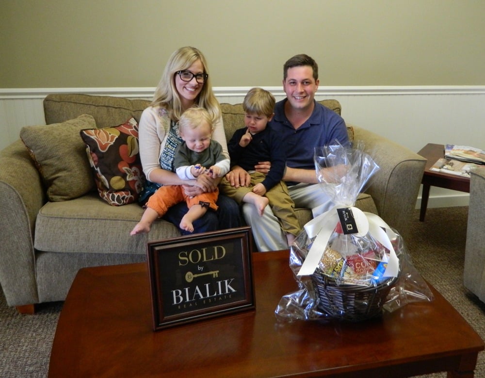 Family of four celebrating a home closing on a sofa beside a SOLD sign and gift basket.