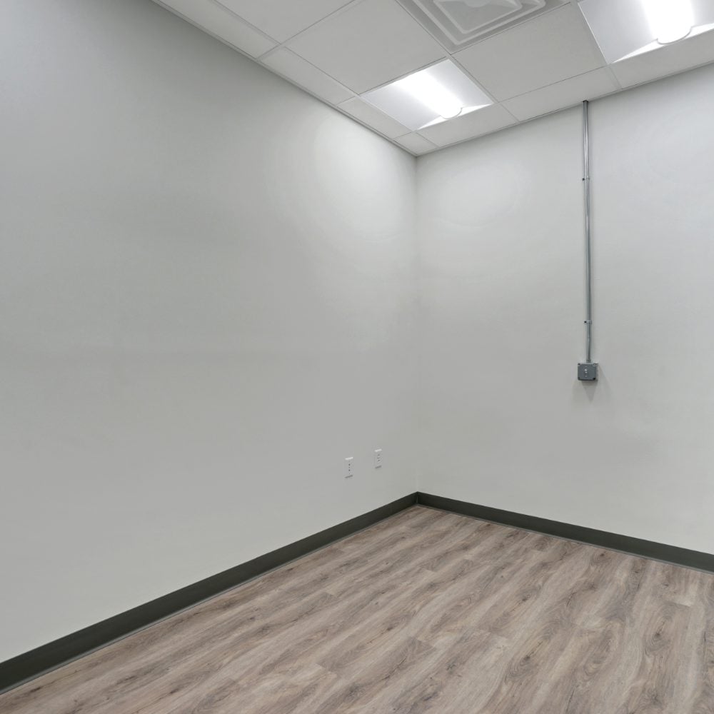 Vacant room with wood-look flooring, white walls, and a suspended ceiling with fluorescent lights.