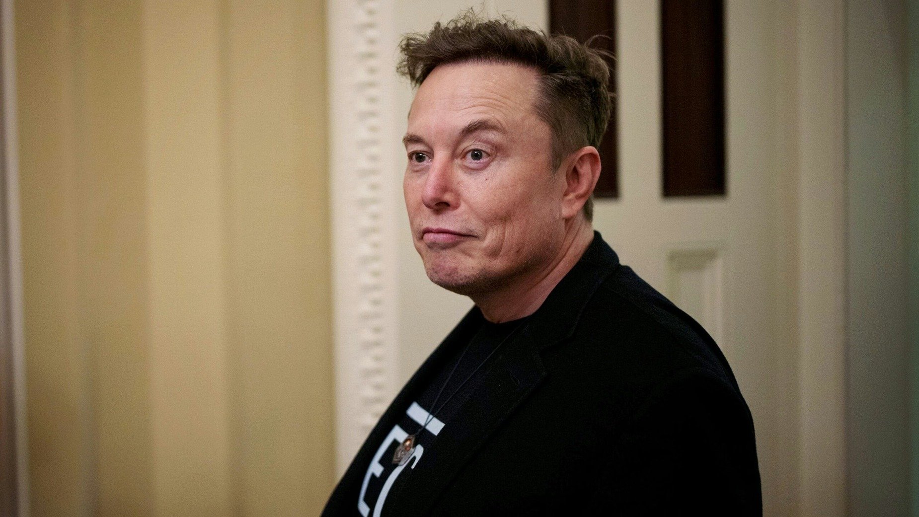 Elon Musk in a black jacket and graphic T-shirt, looking to the side.