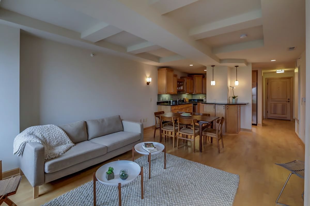 Open-plan living room with a gray sofa, dining table, and warm wood kitchen cabinetry.