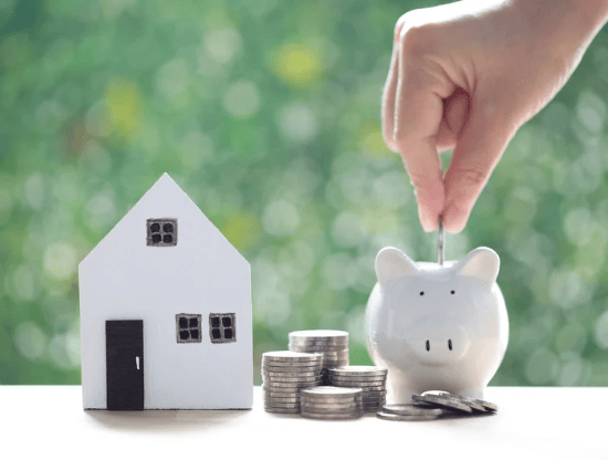 A hand drops a coin into a piggy bank beside a model house and coins.