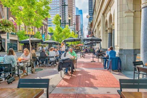 This lively scene captures a sunny day in downtown Calgary, Alberta, where people enjoy food and drinks on a bustling outdoor patio. Surrounded by tall buildings and shaded by umbrellas, the relaxed atmosphere blends urban energy with casual comfort. The street is lined with trees and historic architecture, contributing to the city's inviting and walkable vibe.
