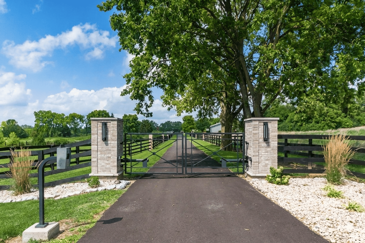 Gated entry drive at a luxury estate property in Delaware County Ohio with custom ironwork, brick pillars, board fencing, and mature tree canopy