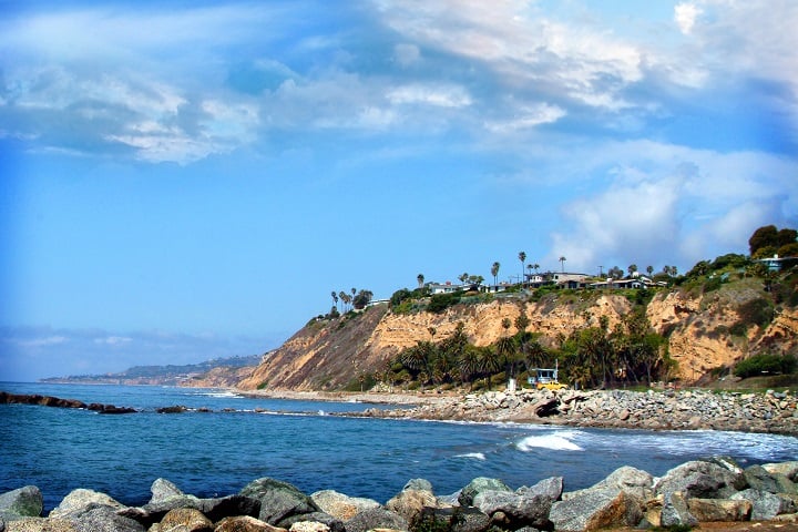 Rocky shoreline beneath coastal cliffs topped with homes and palm trees.
