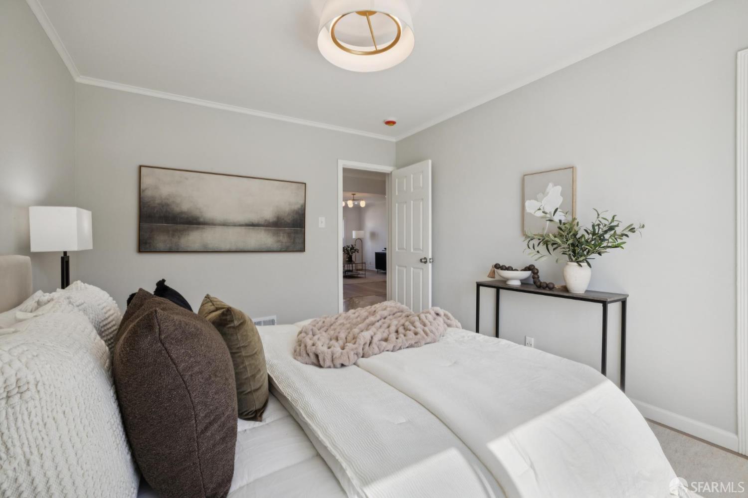 Staged bedroom with a white bed, neutral pillows, abstract wall art, and a console table with greenery.
