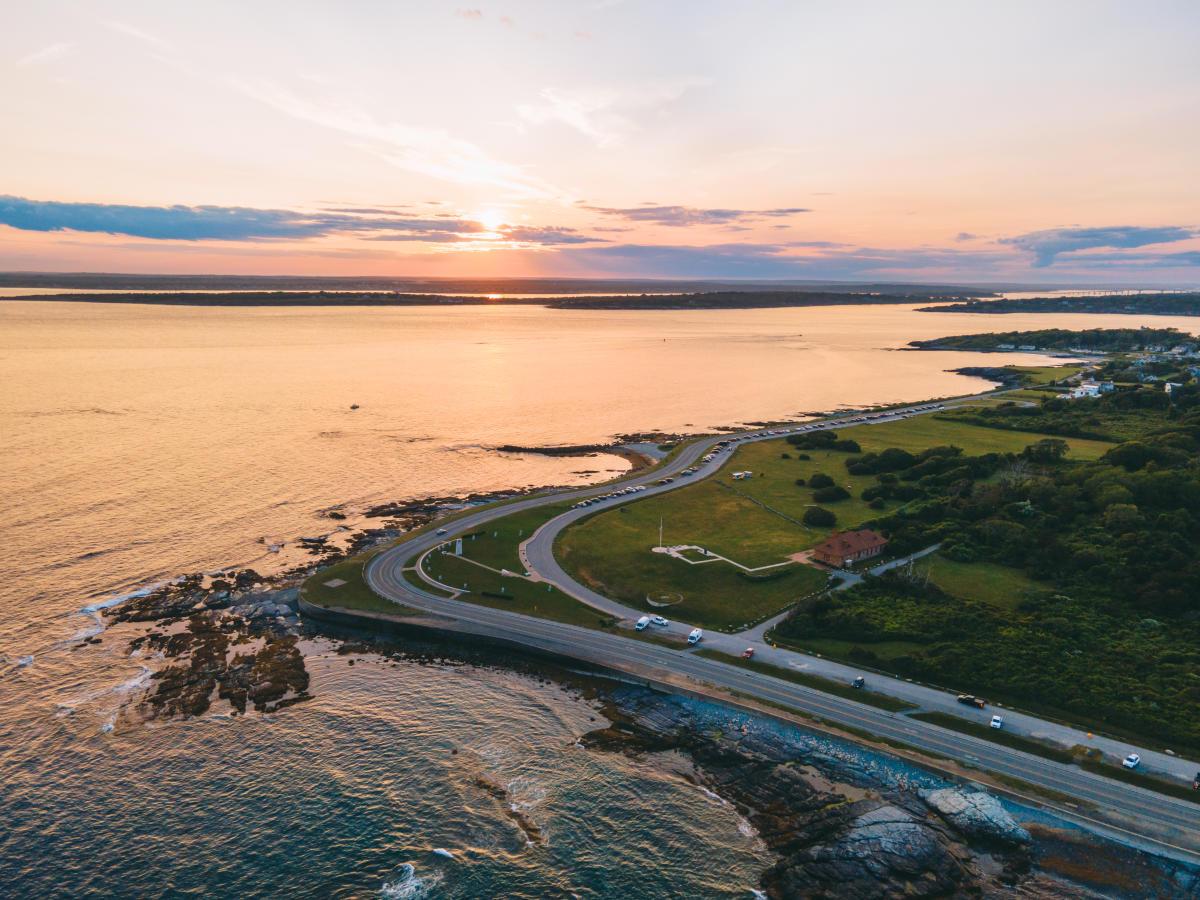 Aerial sunset over a coastal road curving around a grassy, rocky shoreline.