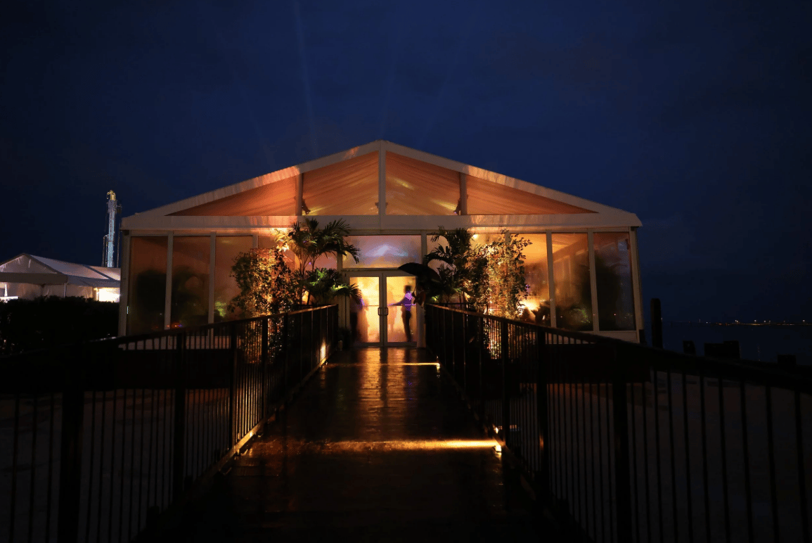 Illuminated glass pavilion at night, reached by a walkway beside the water.