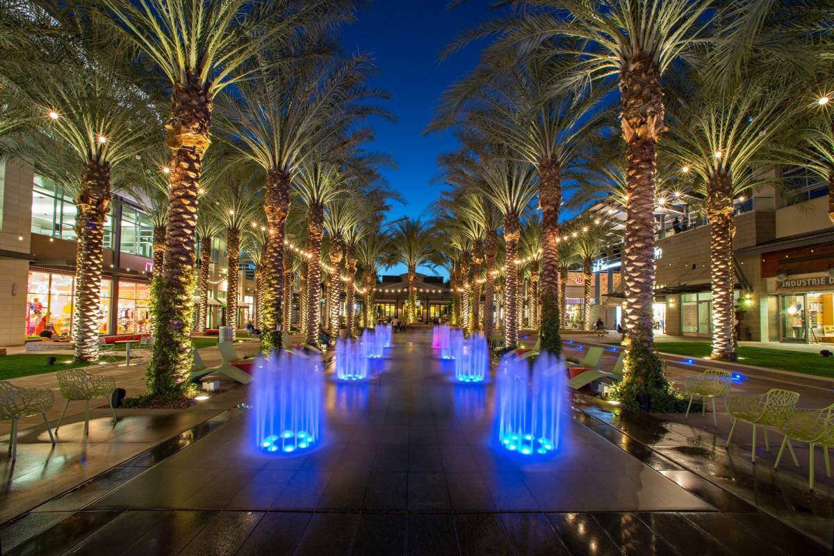 Outdoor shopping plaza with illuminated palm trees and blue-lit fountains.