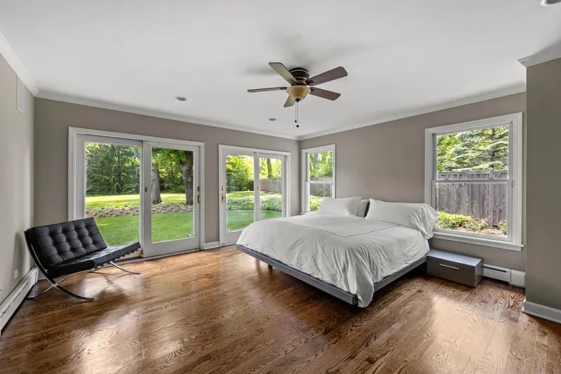 Bedroom with a white bed, hardwood floors, and large windows overlooking a green yard.