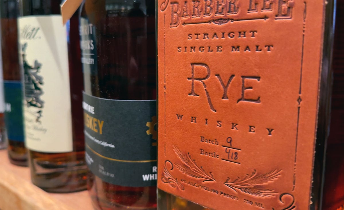 A close-up of a whiskey bottle labeled "Barber Lee Rye Whiskey," with other bottles blurred in the background.