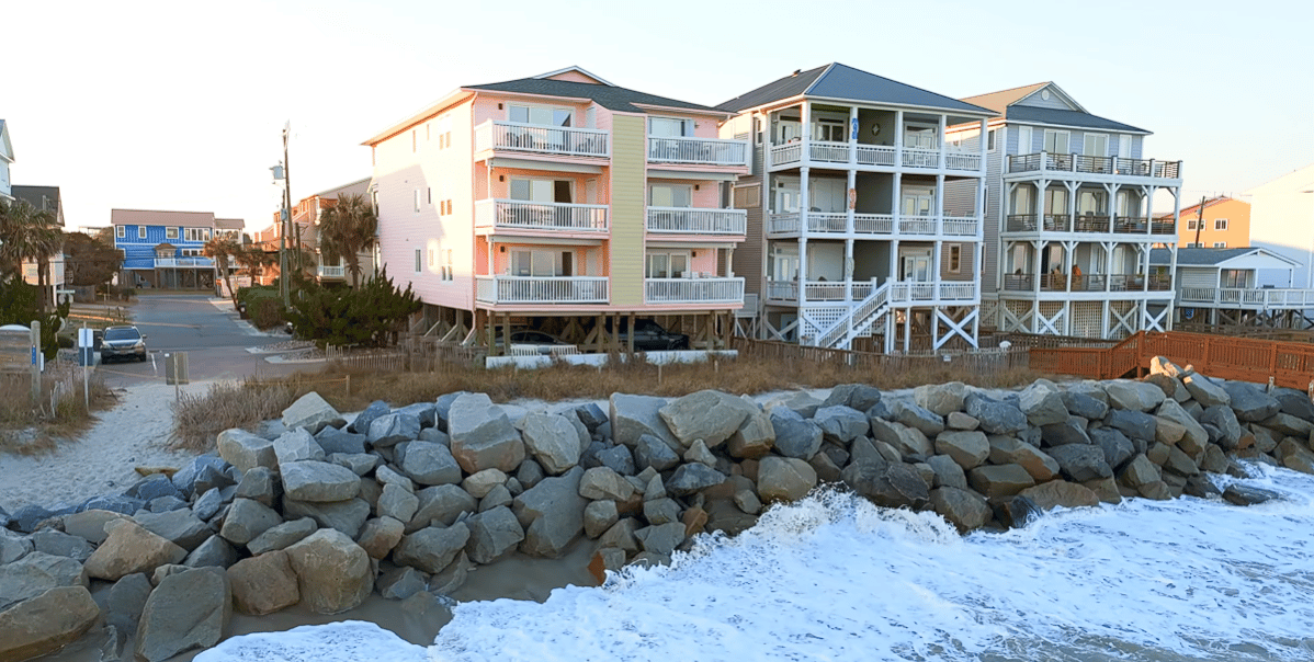 Oceanfront condos with stacked balconies behind a rocky seawall and breaking waves.
