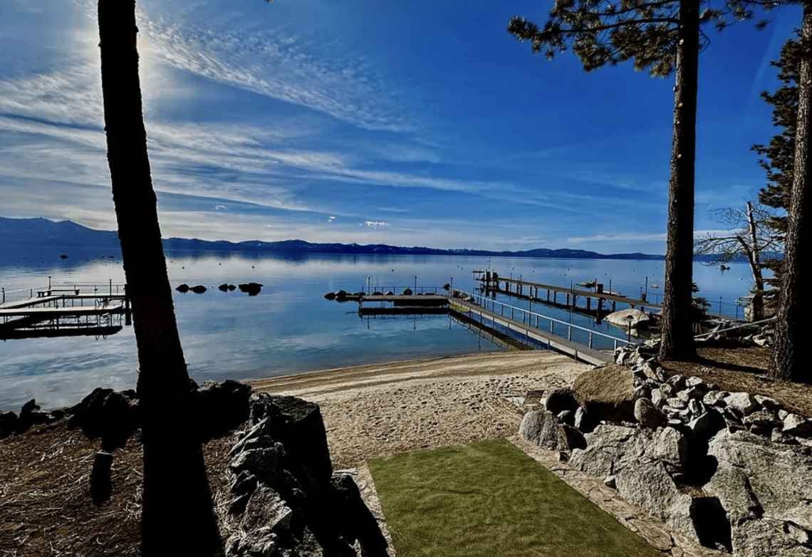 Lakefront beach and docks beside calm blue water, framed by pine trees and rocks.