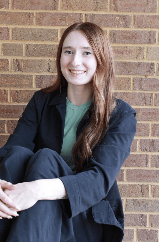 Ariel Smith seated against a brick wall, wearing a dark jacket and green shirt.
