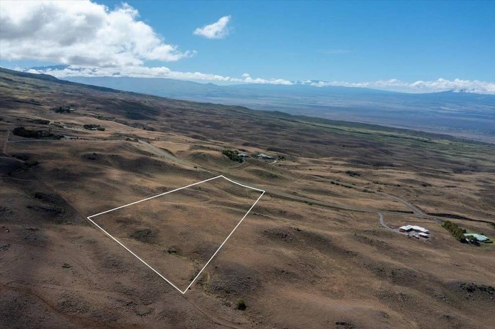 Aerial view of a white-outlined rural lot across dry rolling terrain, with distant mountains and a winding road.