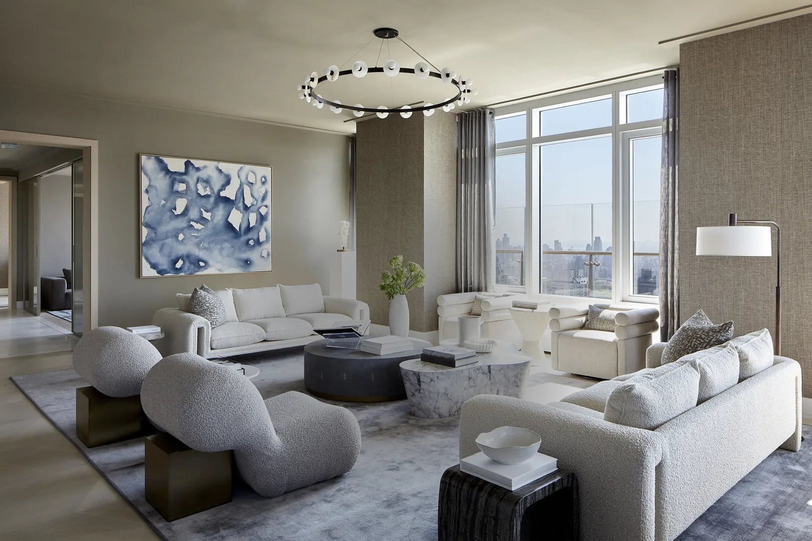 Living room with pale sculptural seating, a circular chandelier, and city skyline views through tall windows.