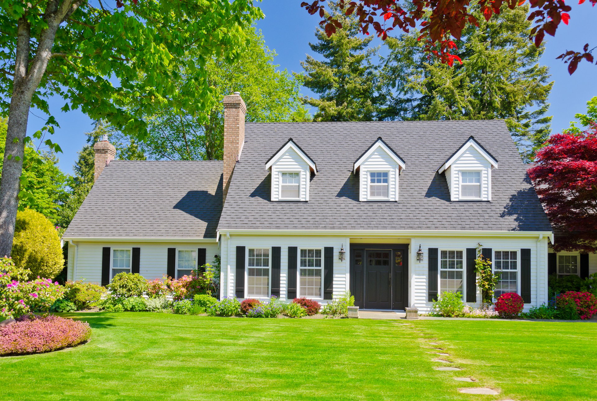 White two-story home with three roof dormers, black shutters, colorful landscaping, and a broad green lawn.