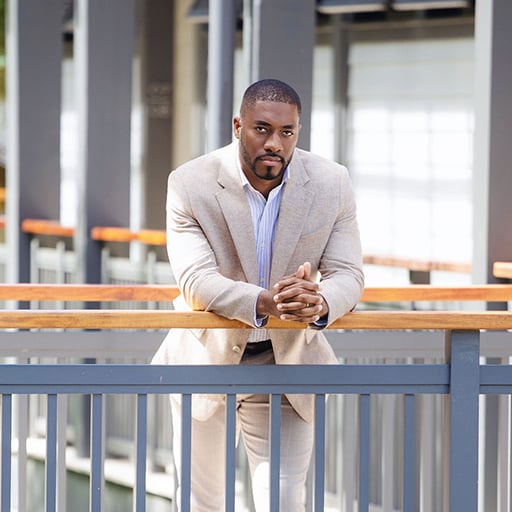 Man in a beige suit leaning on a wooden handrail inside a bright modern building.