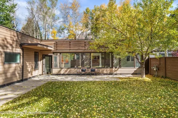 Contemporary single-story home with a broad wall of windows, patio chairs, and autumn leaves covering the lawn.