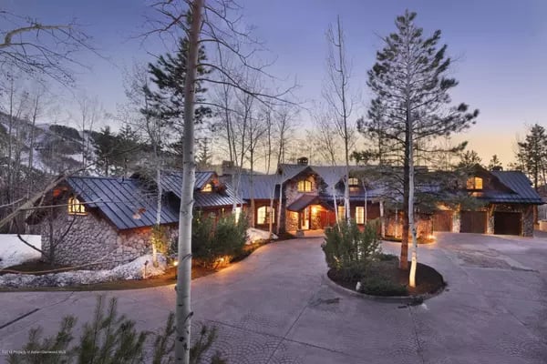 Stone-and-timber home with a metal roof, warmly lit windows, and a curved driveway at dusk.