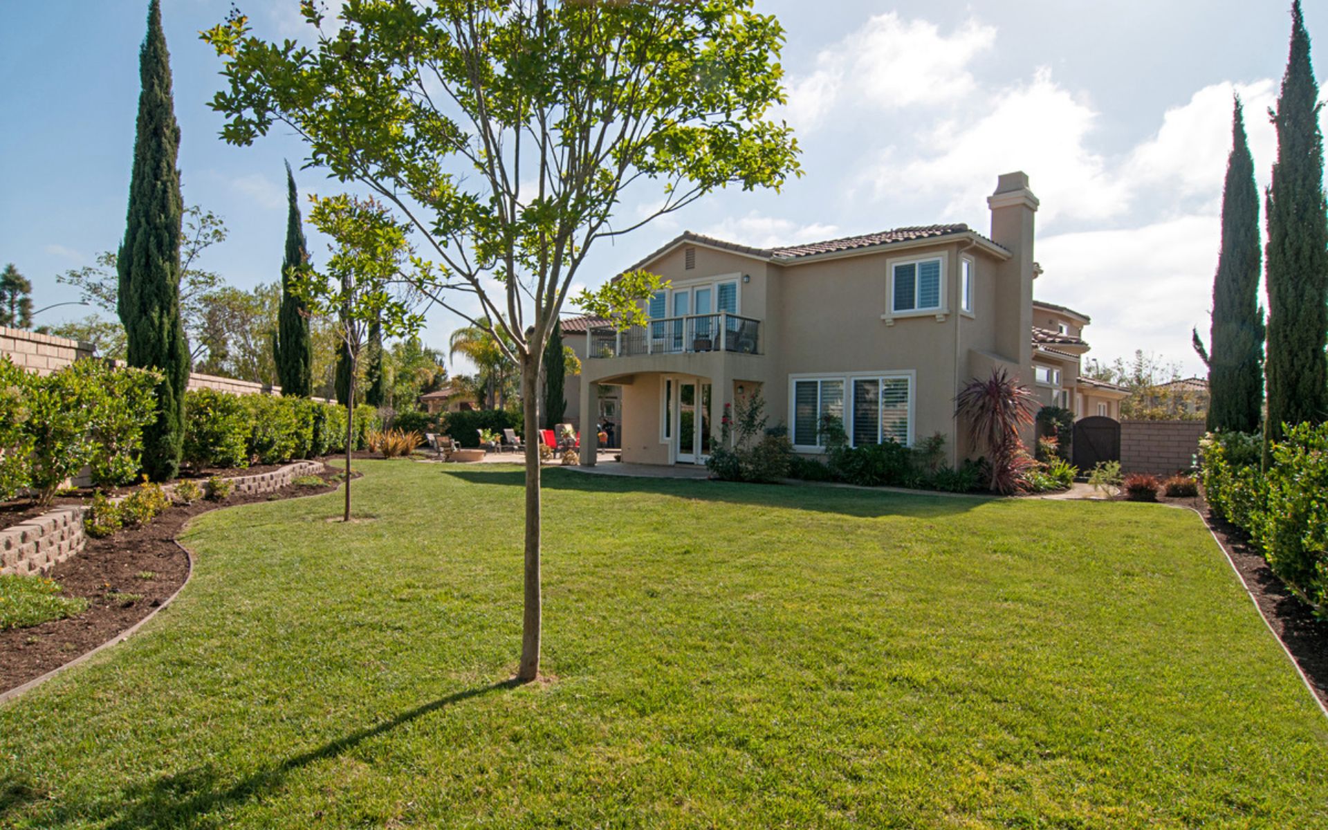 Two-story stucco home overlooking a broad landscaped backyard with a lawn, trees, and upper balcony.