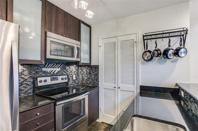 Compact kitchen with dark wood cabinets, stainless appliances, mosaic backsplash, hanging pots, and louvered pantry doors.