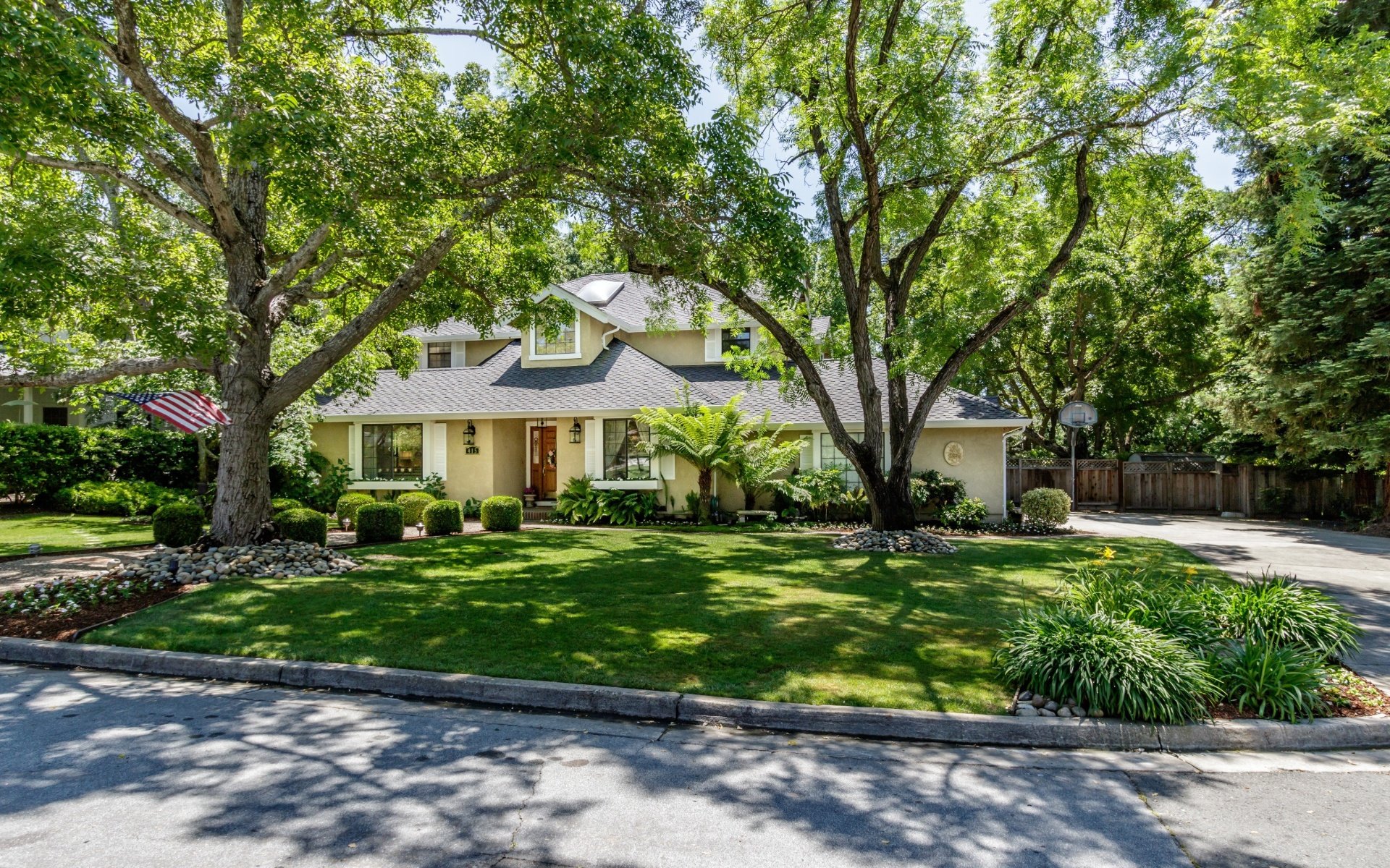 Two-story cream house framed by mature trees, with a landscaped lawn and driveway.