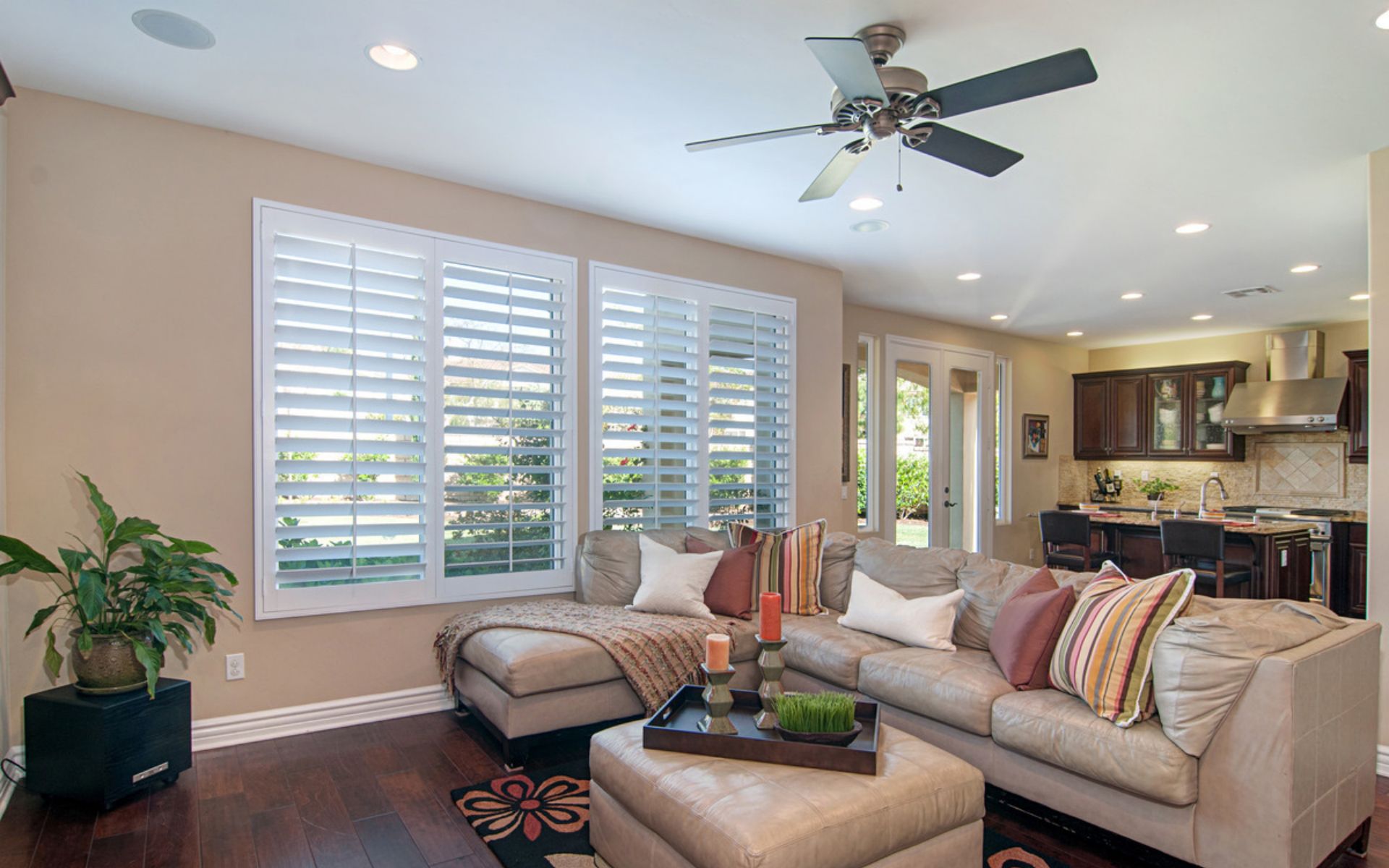 Living room with a beige sectional, plantation shutters, and an open view into the kitchen.