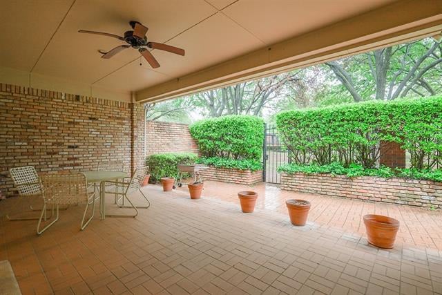 Covered brick patio with terracotta tile, ceiling fan, outdoor seating, and potted plants beside green hedges.