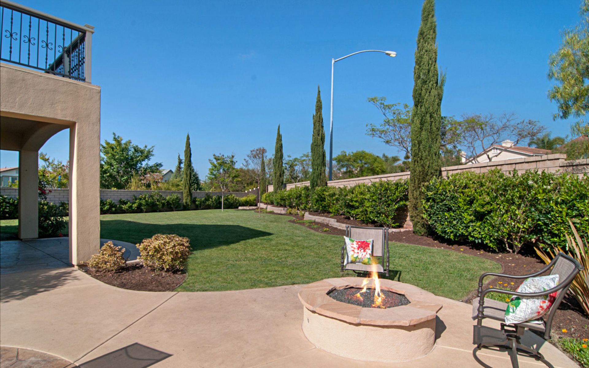 Backyard patio with a round fire pit, two chairs, and a lawn bordered by tall hedges.