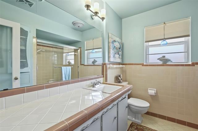 Bathroom with a tiled vanity, glass-enclosed shower, and two windows with blinds.