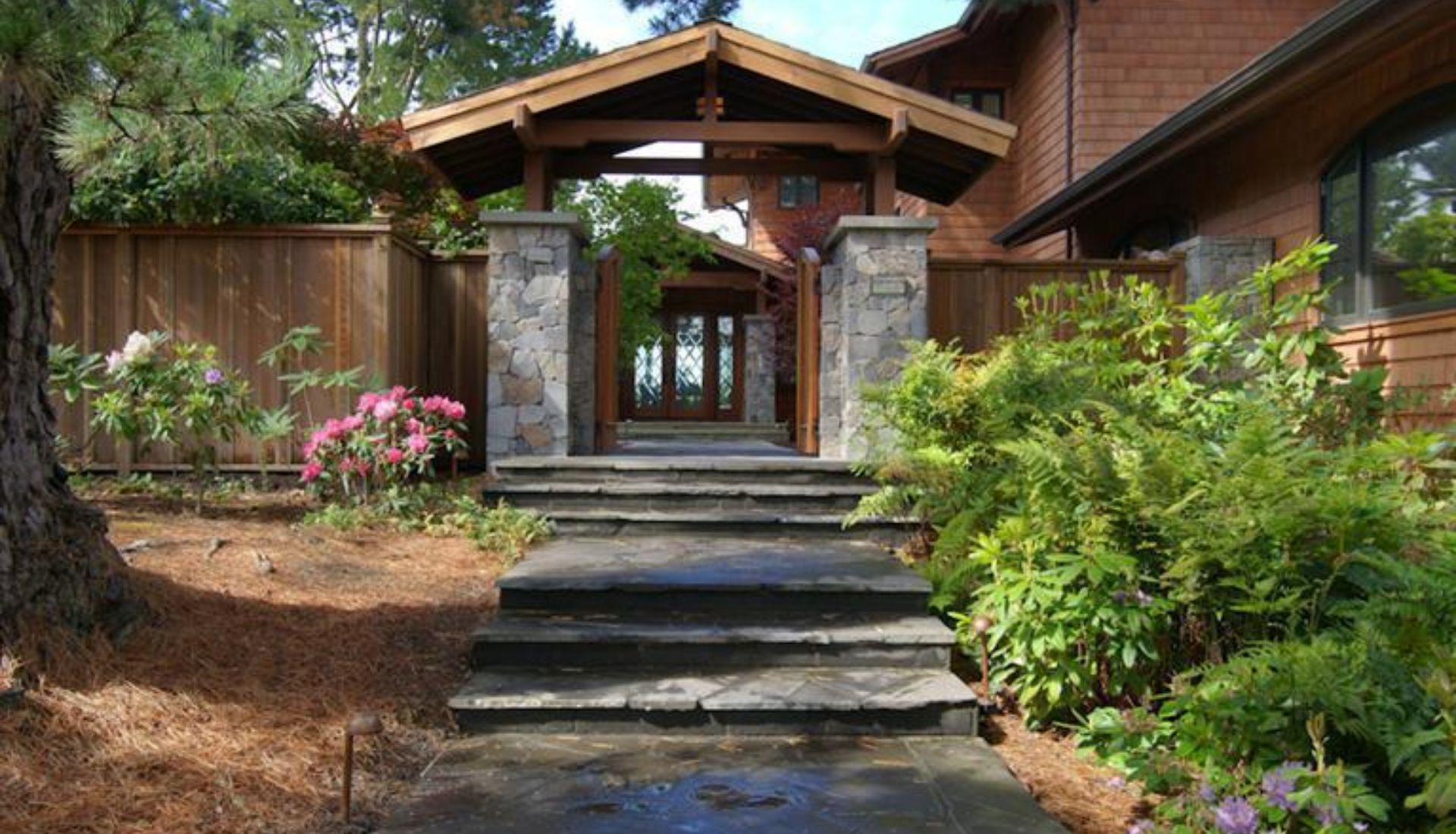 Home entrance with broad stone steps, stone pillars, wooden fencing, and flowering shrubs.