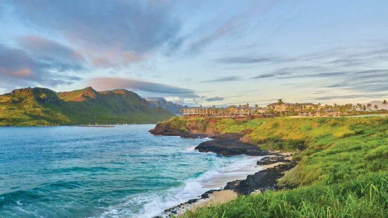Coastal resort above a rocky beach, with turquoise water and mountains across the bay.